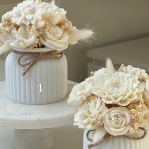 May include: Two white, ribbed vases with floral arrangements. The flowers are cream-colored and include roses and other blooms. The vases are tied with twine. One vase is on a marble cake stand.