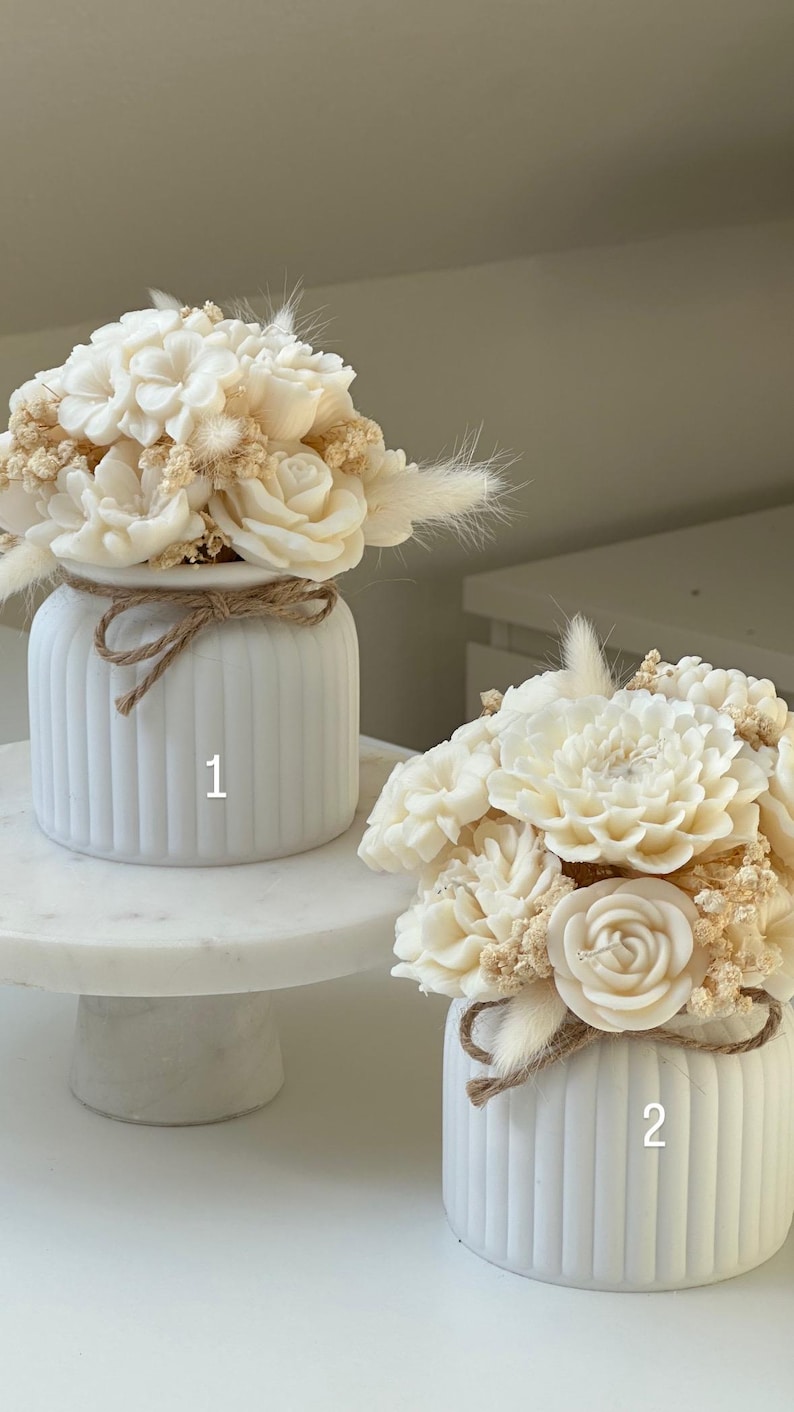 May include: Two white, ribbed vases with floral arrangements. The flowers are cream-colored and include roses and other blooms. The vases are tied with twine. One vase is on a marble cake stand.