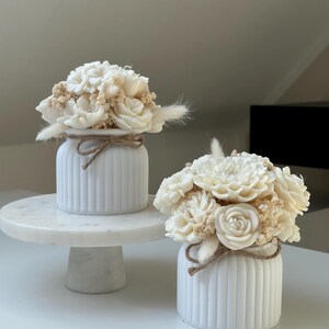 May include: Two white, ribbed ceramic vases, each adorned with a bouquet of cream-colored flowers and dried accents. The vases are tied with twine. One vase sits on a white marble cake stand.
