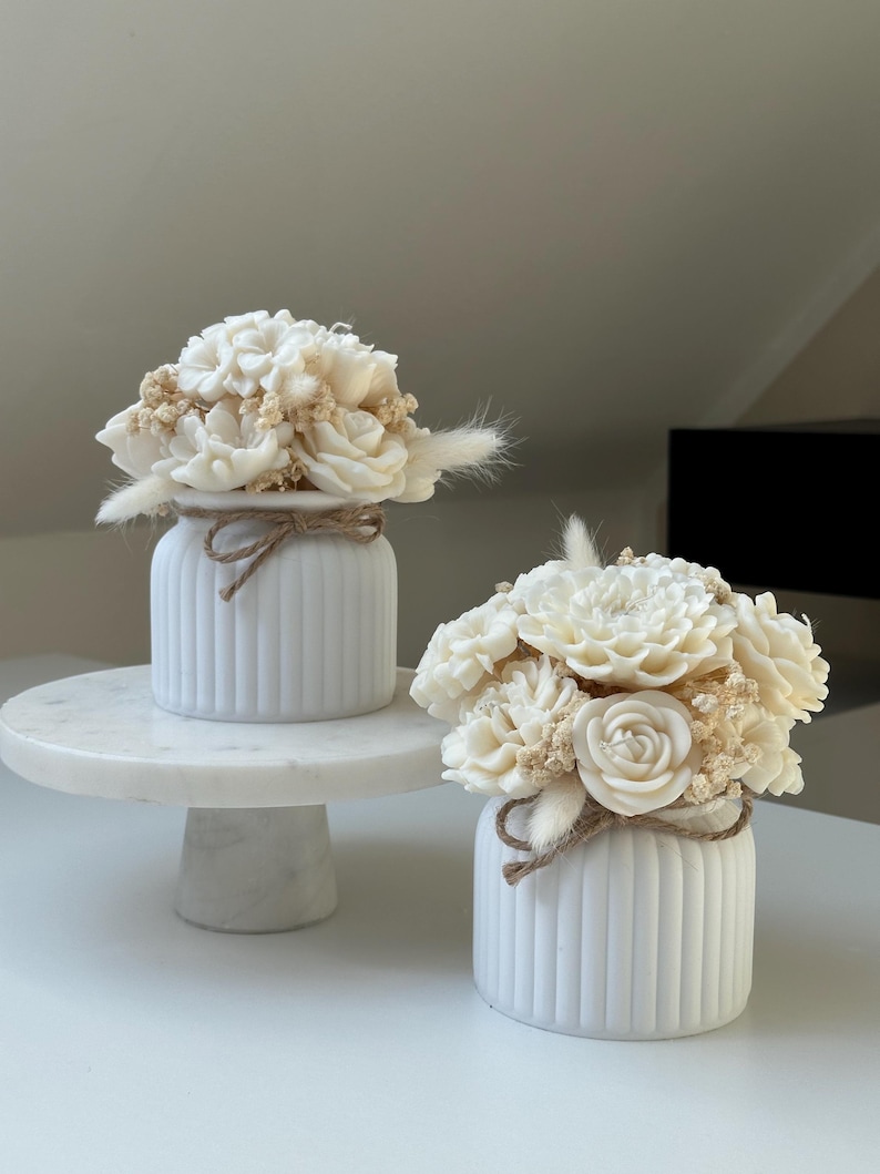 May include: Two white, ribbed ceramic vases, each adorned with a bouquet of cream-colored flowers and dried accents. The vases are tied with twine. One vase sits on a white marble cake stand.