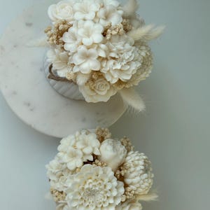 May include: Two ivory-colored floral arrangements. The top arrangement is in a white vase with a rope detail, and the bottom arrangement is on a white surface. Both arrangements feature a variety of flowers and dried accents.