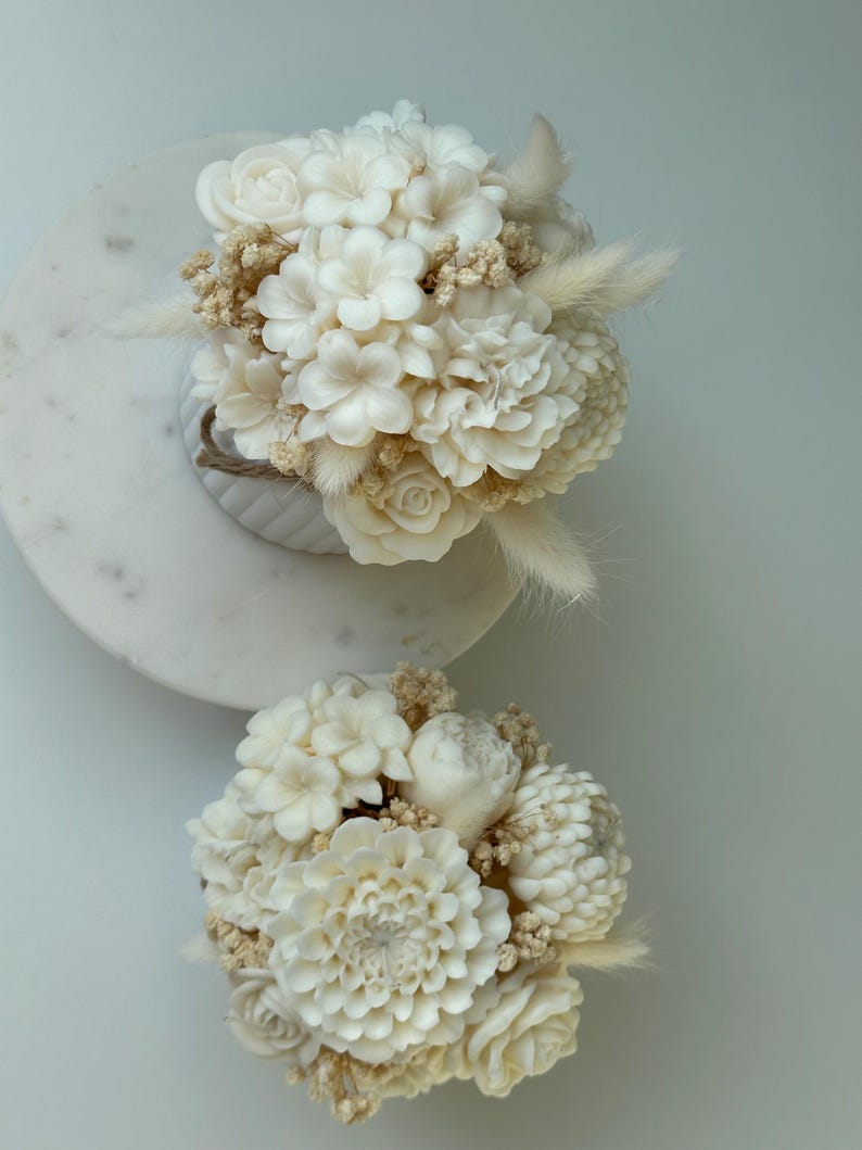 May include: Two ivory-colored floral arrangements. The top arrangement is in a white vase with a rope detail, and the bottom arrangement is on a white surface. Both arrangements feature a variety of flowers and dried accents.