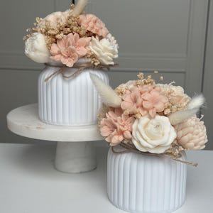 May include: Two white, ribbed ceramic vases, each holding a floral arrangement. The arrangements feature peach, cream, and tan-colored flowers, including roses and peonies. The vases are tied with twine and one sits on a marble cake stand.