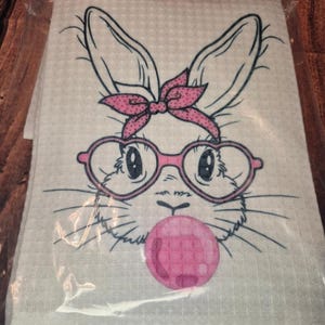 Puede incluir: Una pila de toallas blancas de tejido de gofre con la cara de un conejito de dibujos animados. El conejito tiene gafas rosas, un lazo rosa y una burbuja de chicle rosa. La cara del conejito está delineada en negro.