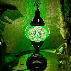 May include: A green mosaic table lamp with a white floral design. The lamp has a round base and a decorative finial on top. The lamp is lit and casting a warm glow.