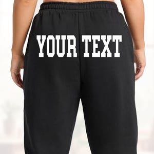 May include: Black sweatpants with the text "YOUR TEXT" in white block letters across the back. The pants have a drawstring waistband and a relaxed fit. The image is taken from the back.