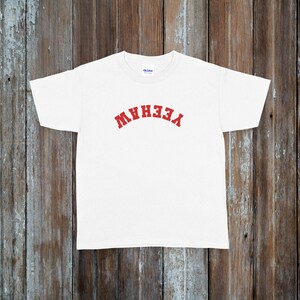 May include: White t-shirt with the word "YEEHAW" in red, arched lettering across the chest. The shirt is made of a soft material and is displayed on a rustic wooden background.