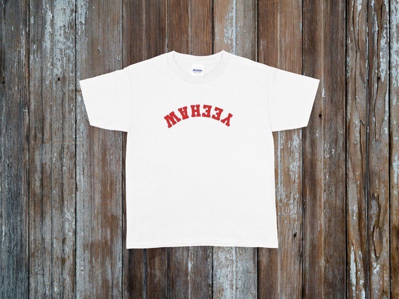 May include: White t-shirt with the word "YEEHAW" in red, arched lettering across the chest. The shirt is made of a soft material and is displayed on a rustic wooden background.