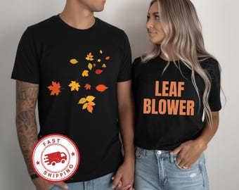 May include: Two black short-sleeved t-shirts. The left shirt features a scattered pattern of orange, yellow, and red autumn leaves. The right shirt features the text LEAF BLOWER in bold orange capital letters centered on the chest.