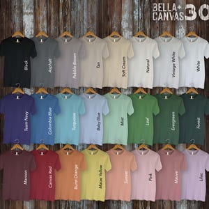 May include: A display of 30 Bella + Canvas 3001 t-shirts in various colors, including black, white, navy, red, orange, yellow, green, blue, and brown. The shirts are arranged in rows on a wooden background.