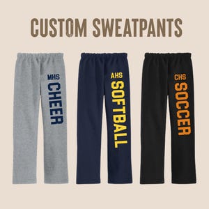 May include: Three pairs of custom sweatpants are displayed. The first pair is gray with "MHS CHEER" in navy blue. The second pair is navy blue with "AHS SOFTBALL" in yellow. The third pair is black with "CHS SOCCER" in orange.