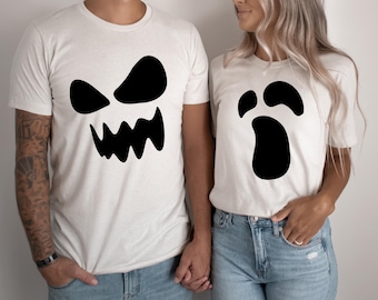 May include: Two white short-sleeved t-shirts featuring black graphic prints. The left shirt displays a jagged, angry ghost face with sharp teeth, while the right shirt features a simple, rounded ghost face with wide eyes and an open mouth.