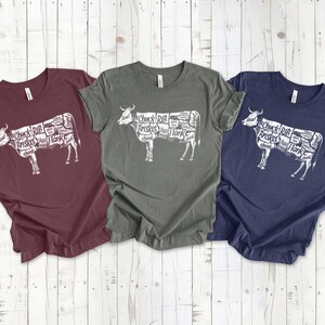 May include: Three t-shirts with a white graphic of a cow with different cuts of meat labeled. The shirts are maroon, dark green, and navy blue.