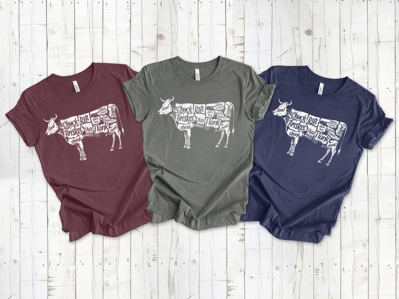 May include: Three t-shirts with a white graphic of a cow with different cuts of meat labeled. The shirts are maroon, dark green, and navy blue.