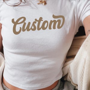 May include: White crop top with a brown "Custom" graphic in a retro font.