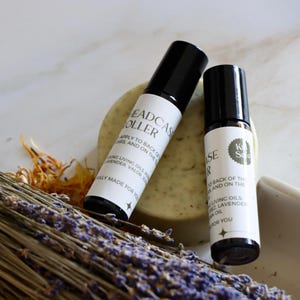 Könnte beinhalten: Zwei kleine Glas-Rollflaschen mit schwarzen Kappen und Etiketten mit der Aufschrift "Headcase Roller" und "Ease" auf weißem Hintergrund mit getrockneter Lavendel und einer Seife.