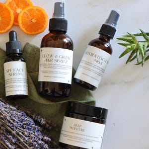 K&ouml;nnte beinhalten: Eine Auswahl an Hautpflegeprodukten in braunen Glasflaschen und -gl&auml;sern, darunter ein Serum, Haarspray, Gesichtsspray und eine Feuchtigkeitscreme. Die Produkte sind mit Orangenscheiben, Lavendel und Gr&uuml;nzeug auf einer wei&szlig;en Oberfl&auml;che angeordnet.