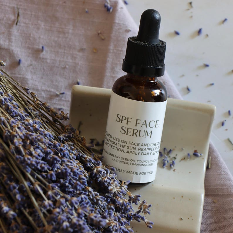 K&ouml;nnte beinhalten: Eine braune Glasflasche mit SPF-Gesichtsserum und einer schwarzen Pipette auf einem wei&szlig;en Keramiktablett. Das Etikett enth&auml;lt Produktinformationen. Ein B&uuml;ndel getrockneter Lavendel und Lavendelknospen befinden sich im Vordergrund.