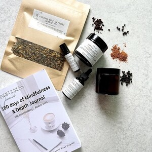 Puede incluir: Una vista cenital presenta una colección de productos de bienestar. Una bolsa de papel marrón de té de hierbas se encuentra junto a un diario titulado "180 días de Mindfulness & Depth Journal". También se incluyen dos frascos negros, un frasco con rodillo y un frasco cuentagotas.