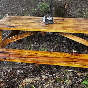 Pode incluir: Uma mesa de piquenique rústica de madeira com bancos anexos. A mesa é feita de madeira castanha clara com grãos e nós visíveis. Um boné de beisebol preto e branco está sobre a mesa. A mesa está ao ar livre.