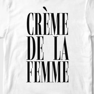 May include: White t-shirt with the text "CR&Egrave;ME DE LA FEMME" in bold, black capital letters. The text is vertically stacked. The shirt has short sleeves and a crew neck.