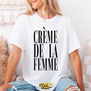 May include: White t-shirt with the text "CR&Egrave;ME DE LA FEMME" in bold black letters. The shirt has short sleeves and is worn by a person. The Pixel Craft logo is at the bottom. The person is wearing blue jeans.