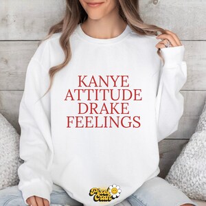 Puede incluir: Sudadera blanca con la frase "Kanye Attitude Drake Feelings" en letras rojas. La prenda tiene cuello redondo y mangas largas. El logotipo de Pixel Craft es visible en la parte inferior.