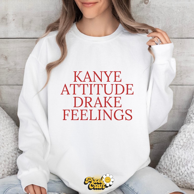 Puede incluir: Sudadera blanca con la frase "Kanye Attitude Drake Feelings" en letras rojas. La prenda tiene cuello redondo y mangas largas. El logotipo de Pixel Craft es visible en la parte inferior.