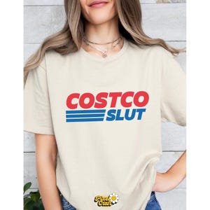 Op de afbeelding: Cr&egrave;mekleurig T-shirt met de woorden "COSTCO SLUT" in rood en blauw. De tekst is in een vetgedrukte, schreefloze lettertype, dat een bekend merklogo imiteert. Het shirt is een casual stijl met korte mouwen.