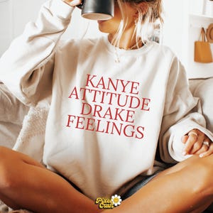 Puede incluir: Sudadera color crema con la frase "Kanye Attitude Drake Feelings" en letras rojas. La sudadera, de estilo casual, es perfecta para el uso diario. La imagen muestra a una persona que lleva la sudadera.