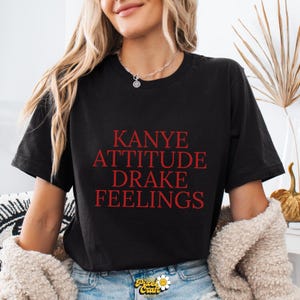 Puede incluir: Camiseta negra con las palabras rojas "KANYE ATTITUDE DRAKE FEELINGS". La camiseta es de cuello redondo y parece estar hecha de un material suave. La camiseta es parte de una l&iacute;nea de ropa.