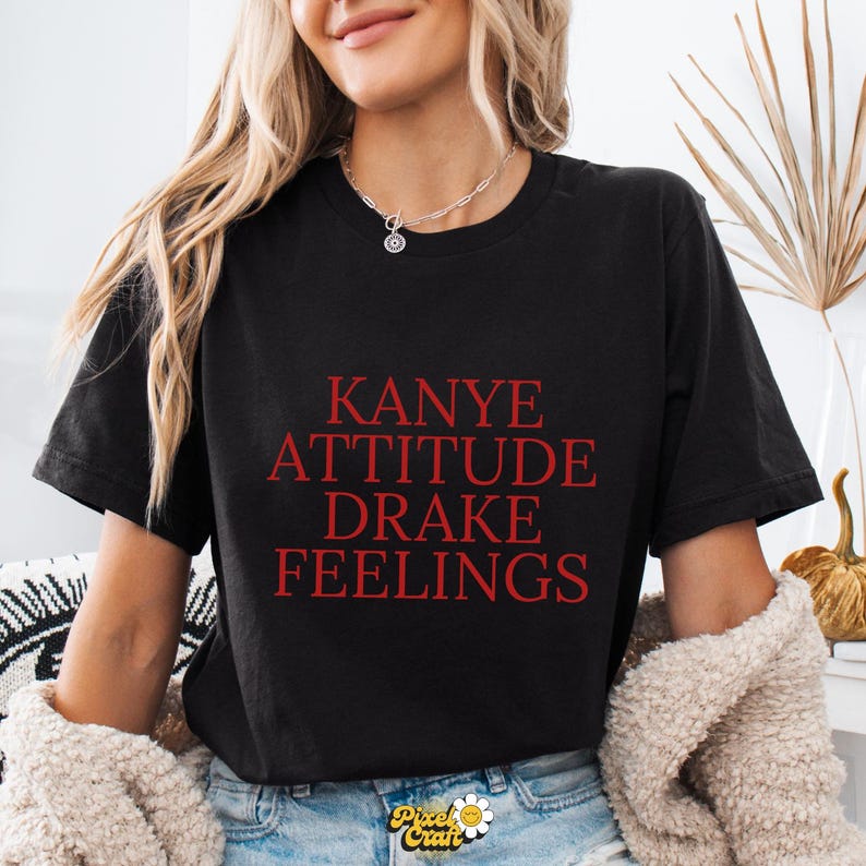 Puede incluir: Camiseta negra con las palabras rojas "KANYE ATTITUDE DRAKE FEELINGS". La camiseta es de cuello redondo y parece estar hecha de un material suave. La camiseta es parte de una l&iacute;nea de ropa.