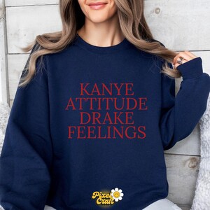 Puede incluir: Sudadera azul marino con las palabras "KANYE ATTITUDE DRAKE FEELINGS" en letras rojas. La sudadera tiene cuello redondo y mangas largas. El logotipo de Pixel Craft est&aacute; en la parte inferior.