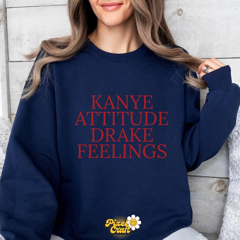 Puede incluir: Sudadera azul marino con las palabras "KANYE ATTITUDE DRAKE FEELINGS" en letras rojas. La sudadera tiene cuello redondo y mangas largas. El logotipo de Pixel Craft est&aacute; en la parte inferior.