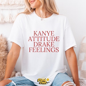 Puede incluir: Camiseta blanca con las palabras "Kanye Attitude Drake Feelings" en letras rojas. La camiseta tiene mangas cortas y cuello redondo. El logotipo de Pixel Craft es visible en la parte inferior.