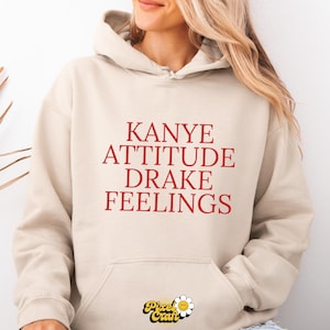 Puede incluir: Sudadera con capucha beige con las palabras "Kanye Attitude Drake Feelings" en letras rojas. La sudadera tiene una capucha con cord&oacute;n y un bolsillo delantero. El logotipo de Pixel Craft est&aacute; en la parte inferior.