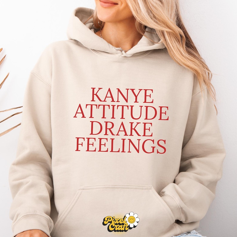 Puede incluir: Sudadera con capucha beige con las palabras "Kanye Attitude Drake Feelings" en letras rojas. La sudadera tiene una capucha con cord&oacute;n y un bolsillo delantero. El logotipo de Pixel Craft est&aacute; en la parte inferior.