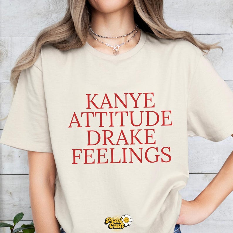 Puede incluir: Camiseta color crema con las palabras rojas "KANYE ATTITUDE DRAKE FEELINGS". La camiseta tiene cuello redondo y mangas cortas. El logo de Pixel Craft est&aacute; en la parte inferior.