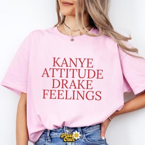 Puede incluir: Camiseta rosa claro con la frase "KANYE ATTITUDE DRAKE FEELINGS" en rojo. La camiseta, de corte holgado, probablemente es de algod&oacute;n o tejido similar. El texto est&aacute; centrado en la parte delantera.