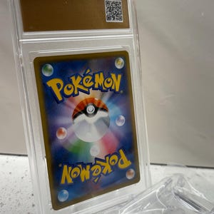 Puede incluir: Una carta coleccionable de Pok&eacute;mon calificada, encerrada en un estuche de pl&aacute;stico transparente. La carta presenta un dise&ntilde;o colorido con la palabra "Pok&eacute;mon" en amarillo. El estuche tiene una parte superior dorada con un c&oacute;digo QR.