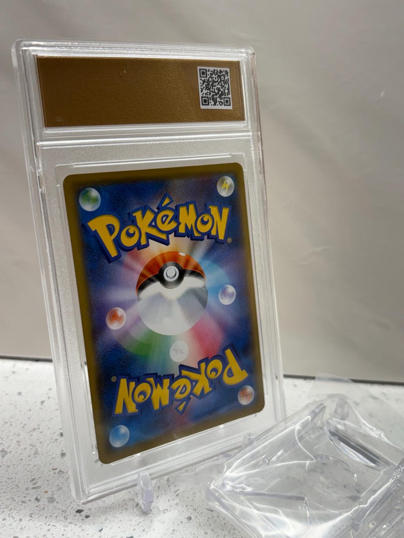 Puede incluir: Una carta coleccionable de Pok&eacute;mon calificada, encerrada en un estuche de pl&aacute;stico transparente. La carta presenta un dise&ntilde;o colorido con la palabra "Pok&eacute;mon" en amarillo. El estuche tiene una parte superior dorada con un c&oacute;digo QR.