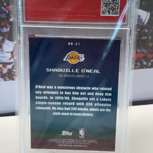 May include: A basketball card featuring Shaquille O'Neal of the Lakers, in a protective case. The card displays the Lakers logo, player details, and career stats. The card is presented on a clear stand.