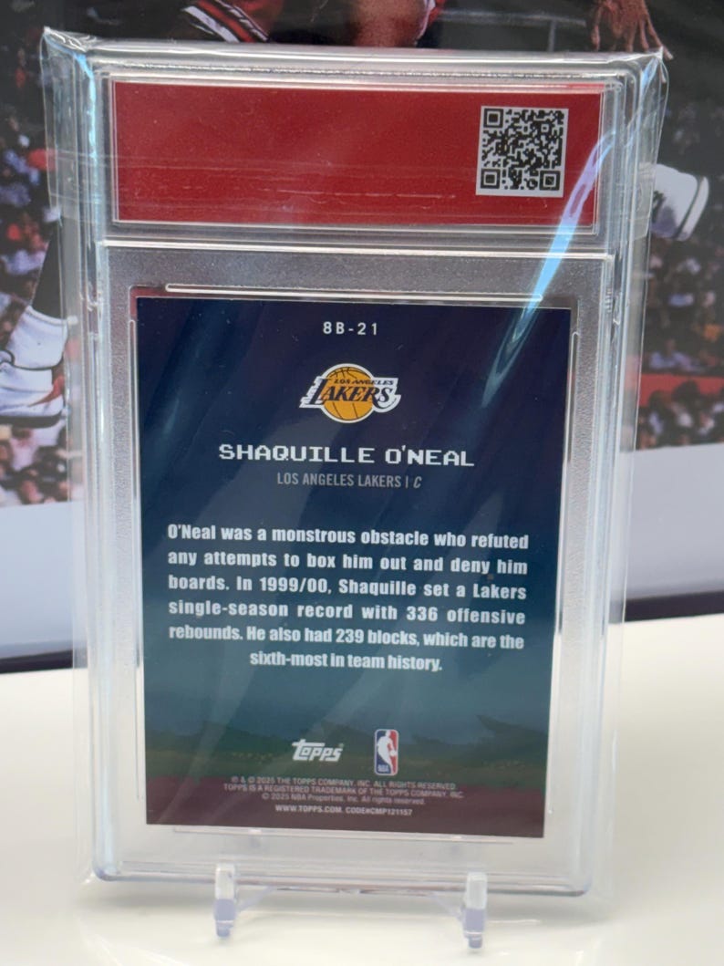 May include: A basketball card featuring Shaquille O'Neal of the Lakers, in a protective case. The card displays the Lakers logo, player details, and career stats. The card is presented on a clear stand.