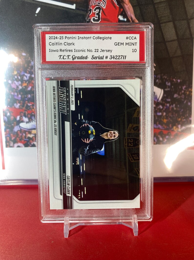 May include: A graded trading card of Caitlin Clark, a basketball player for the Iowa Hawkeyes. The card is from the 2024-25 Panini Instant Collegiate series and is graded GEM MINT 10 by T.C.T. The card features a photo of Clark holding a basketball in a black and white photo. The card is numbered 3422711.