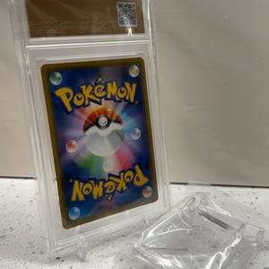 Puede incluir: Una carta coleccionable de Pok&eacute;mon calificada en una funda de pl&aacute;stico transparente. La parte posterior de la carta presenta el logotipo de Pok&eacute;mon en amarillo, con un fondo azul y detalles coloridos. Tambi&eacute;n se ve un soporte y embalaje de pl&aacute;stico transparente.