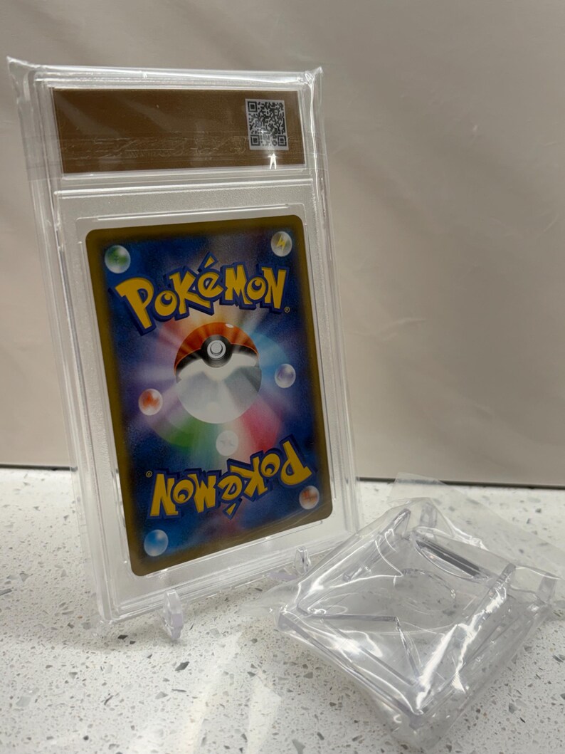 Puede incluir: Una carta coleccionable de Pok&eacute;mon calificada en una funda de pl&aacute;stico transparente. La parte posterior de la carta presenta el logotipo de Pok&eacute;mon en amarillo, con un fondo azul y detalles coloridos. Tambi&eacute;n se ve un soporte y embalaje de pl&aacute;stico transparente.