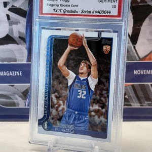 May include: A 2025 Topps NBA Cooper Flagg Flagship Rookie Card, graded GEM MINT 10, encased in a protective holder. The card features a player in a Dallas Mavericks jersey, number 32, shooting a basketball. The card is displayed on a clear stand.