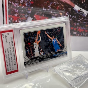 Op de afbeelding: Een basketballkaart met de beoordeling GEM MINT 10, met Victor Wembanyama en Chet Holmgren. De kaart zit in een doorzichtige beschermhoes. De achtergrond toont een wazige afbeelding van een basketballer in actie.