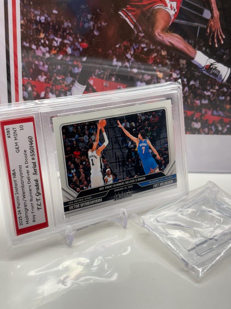 Op de afbeelding: Een basketballkaart met de beoordeling GEM MINT 10, met Victor Wembanyama en Chet Holmgren. De kaart zit in een doorzichtige beschermhoes. De achtergrond toont een wazige afbeelding van een basketballer in actie.