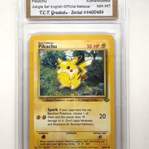 Könnte beinhalten: Eine bewertete 1999 Pokémon Pikachu Karte aus der Jungle Set English Official Release, NM-MT Zustand. Die Karte zeigt eine gelbe Pikachu-Illustration und Text mit "Spark" und "50 HP". Die Karte befindet sich in einer Schutzhülle.